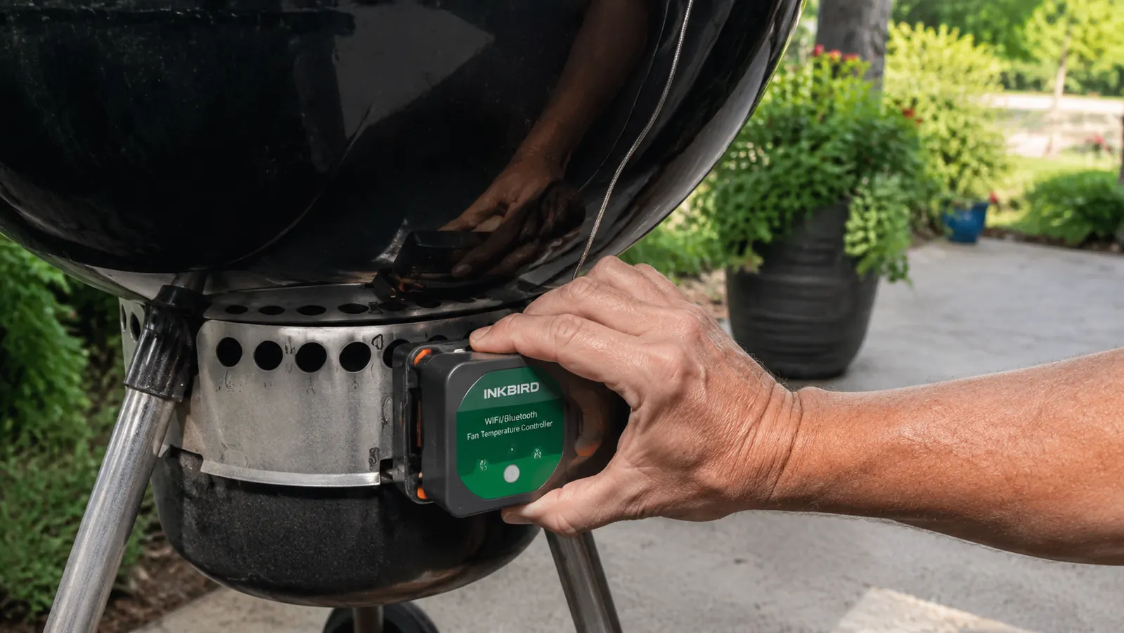 A hand clipping a small fan module onto the lower vent of a kettle grill with a probe wire running toward the lid