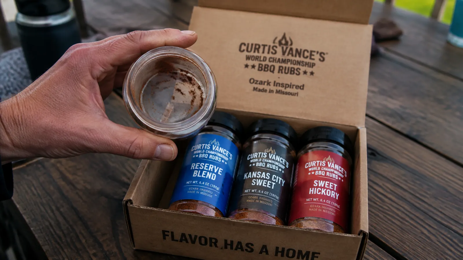 A hand holding an empty small rub jar next to three unopened jars still sealed in the sampler box