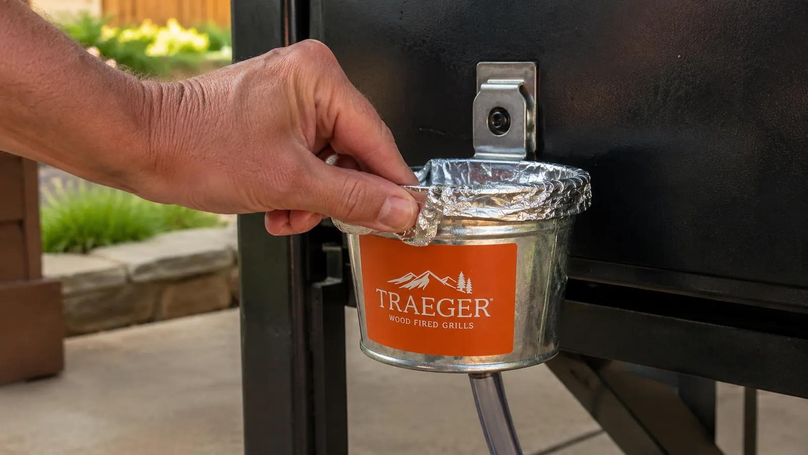 A hand hooking a foil liner bag over the rim of a small metal bucket clipped to the side of a pellet grill