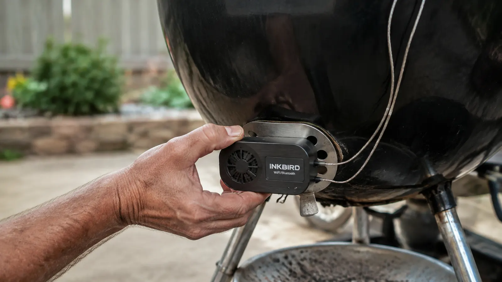 A hand clipping a fan controller module onto the bottom vent of a kettle grill with two probe wires running up toward the lid