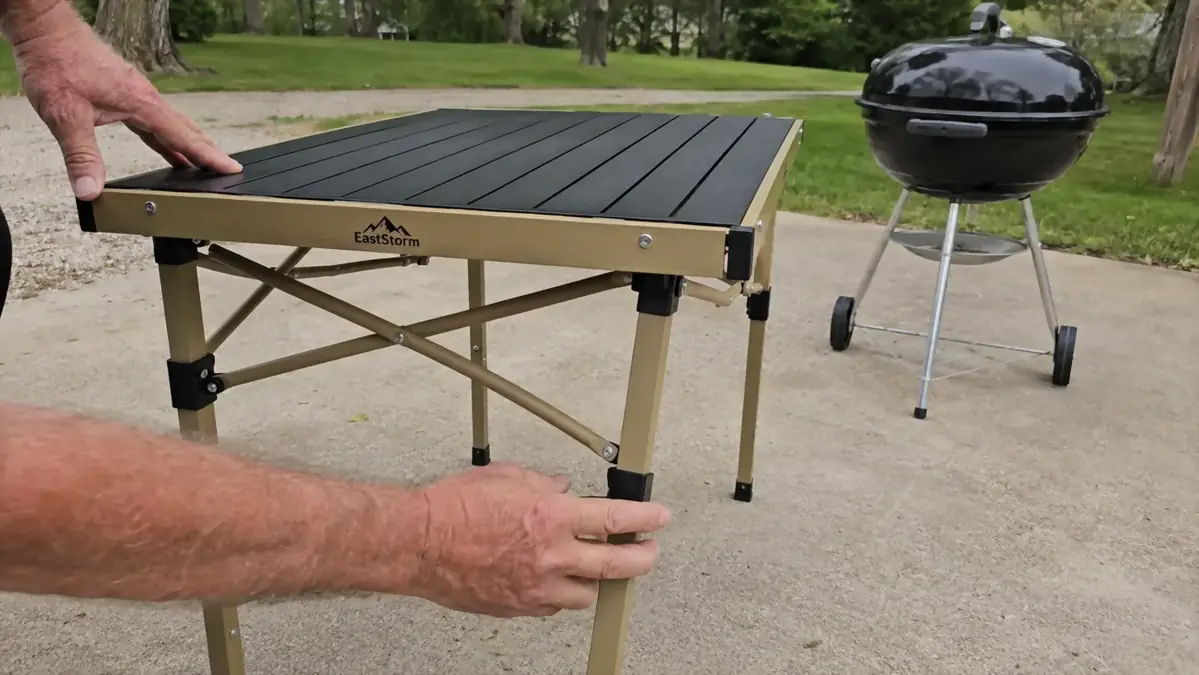Hands setting a folding table down and adjusting its height next to an unlit charcoal grill