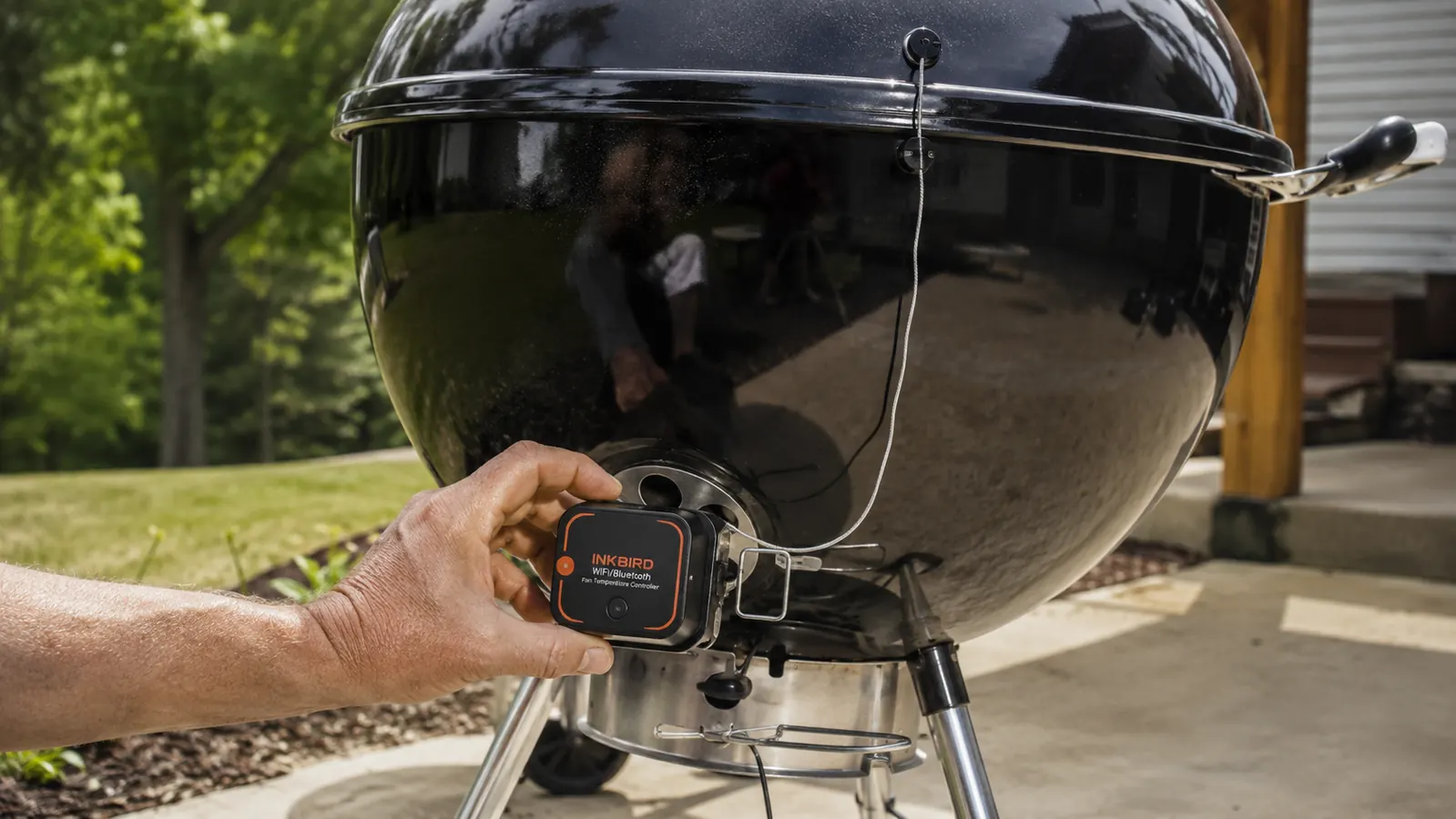 A hand clipping a small fan module onto the bottom vent of a kettle grill with a probe wire running up toward the lid