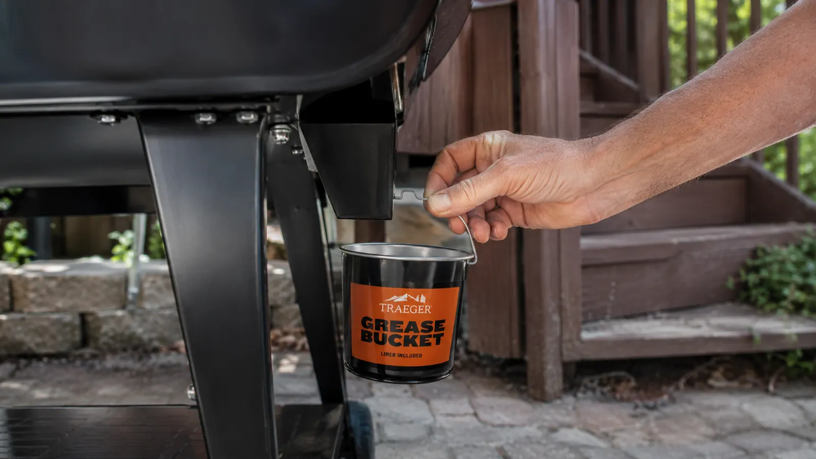 A hand hooking a small metal grease bucket onto a bracket on the side of a pellet grill
