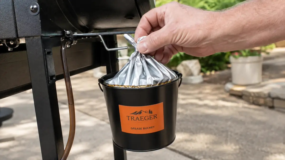 A hand tying off a full foil liner bag pulled from a small bucket on the side of a pellet grill, grease visible along the folded seam