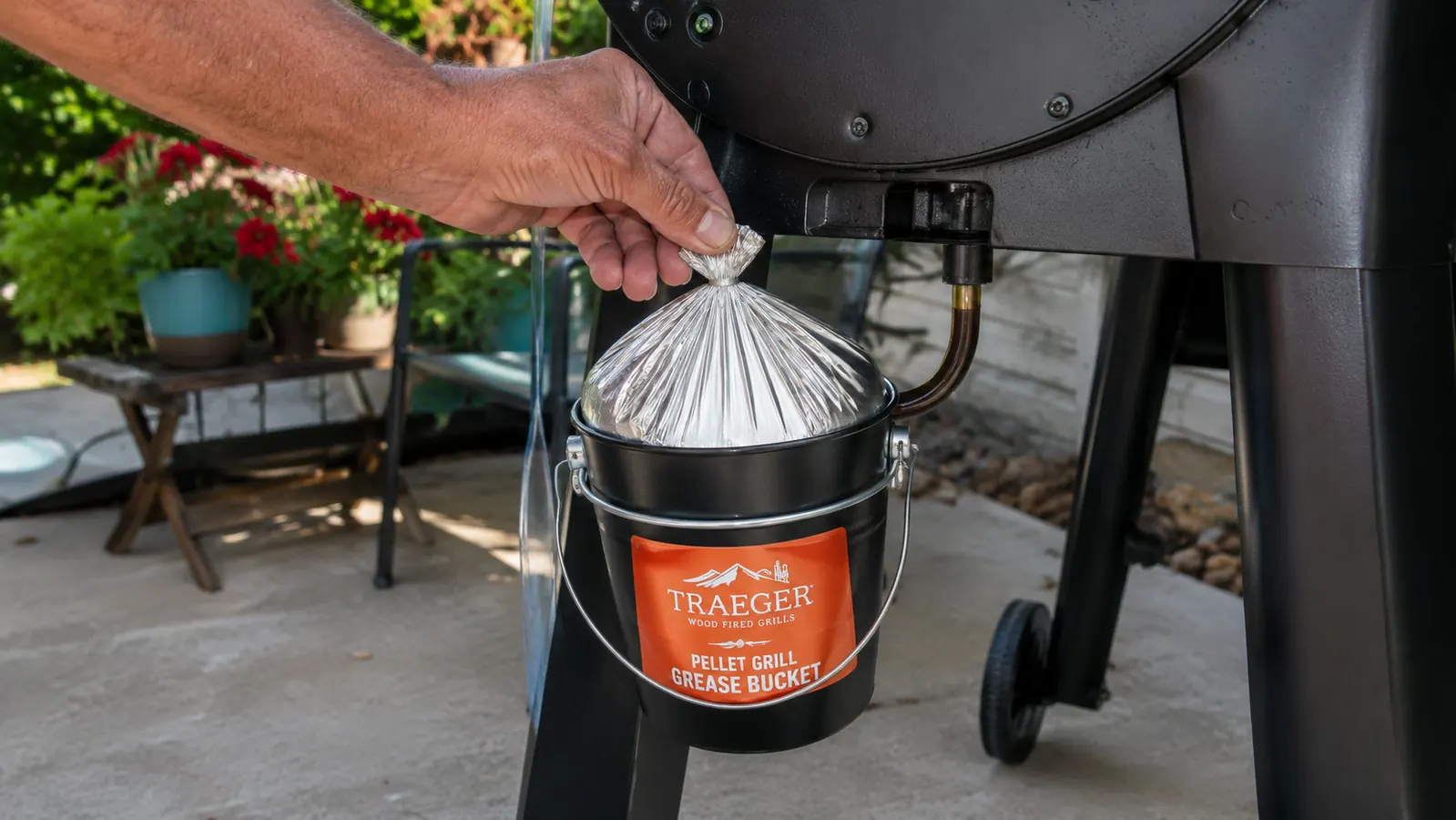 A hand lifting a tied-off foil liner bag out of a small metal bucket clipped to the side of a pellet grill