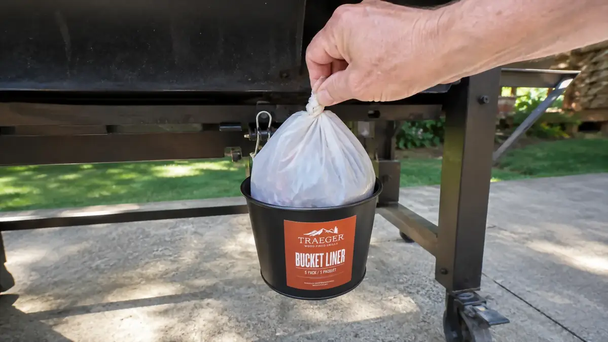 Hand lifting a tied-off liner out of the Traeger grease bucket under a pellet grill